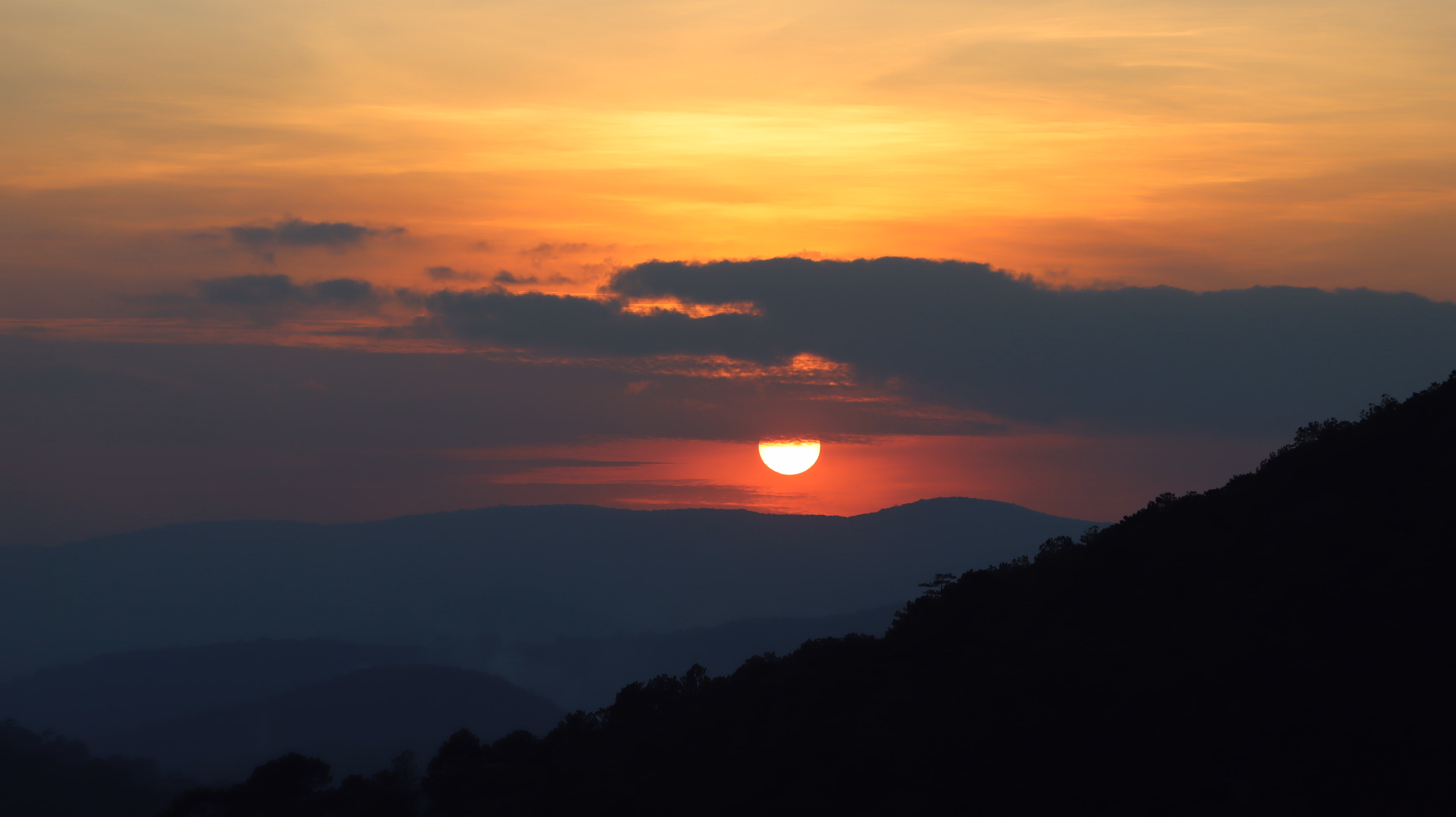 why sunsets look orange and red A dramatic orange and red sunset sky over a silhouette of trees and hills